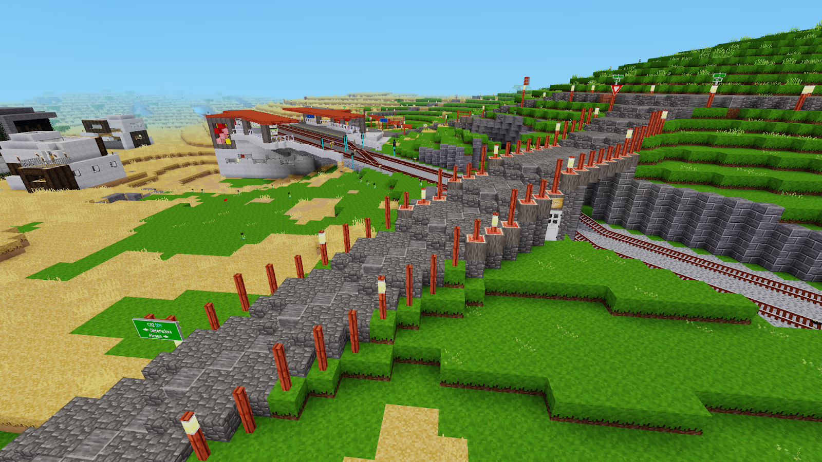 Screenshot of a Minetest world: a stone retaining wall lined with lamp posts curves across green terraced hills toward a railway station, with rail tracks, a road sign and desert village buildings in the distance.