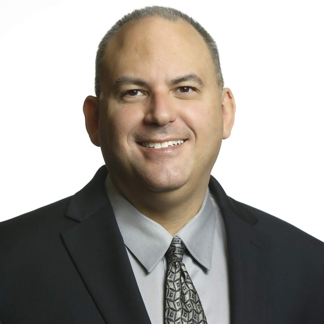 Author photo of Miguel Guhlin, smiling, in a dark suit jacket, gray shirt and patterned tie.
